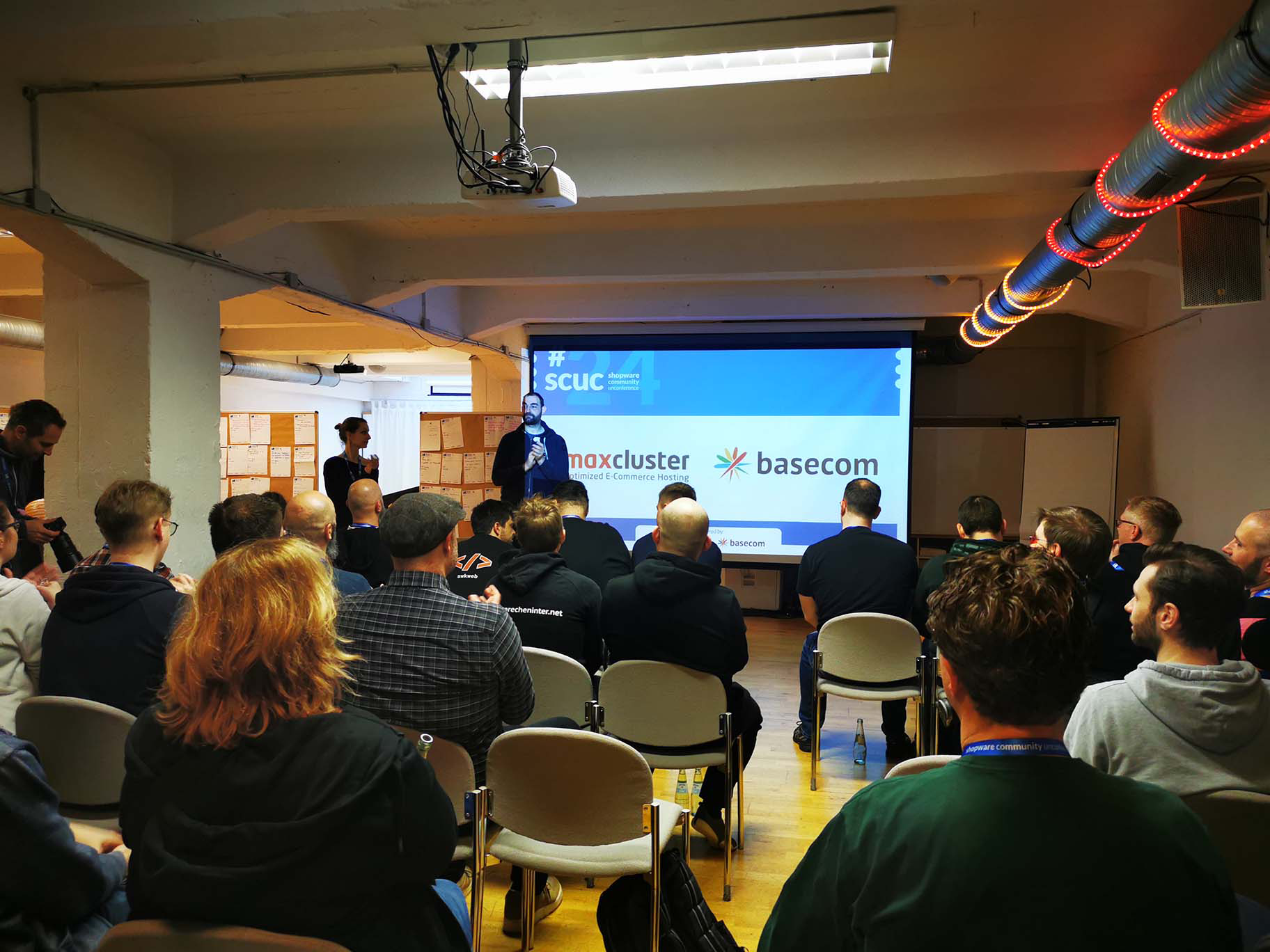 A room full of people, one is in front and talking to the crowd.